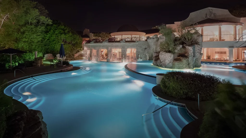 A luxurious, illuminated swimming pool curves around a resort-style building at night, featuring rock formations, lush greenery, and softly glowing underwater lights.
