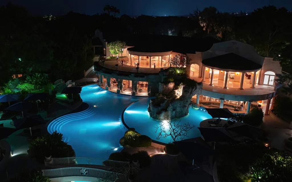 A luxurious, well-lit resort pool at night, featuring curved swim areas, rock formations, and a large building with tall windows and balconies, surrounded by lush greenery and glowing blue water.