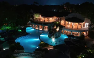 A luxurious, well-lit resort pool at night, featuring curved swim areas, rock formations, and a large building with tall windows and balconies, surrounded by lush greenery and glowing blue water.