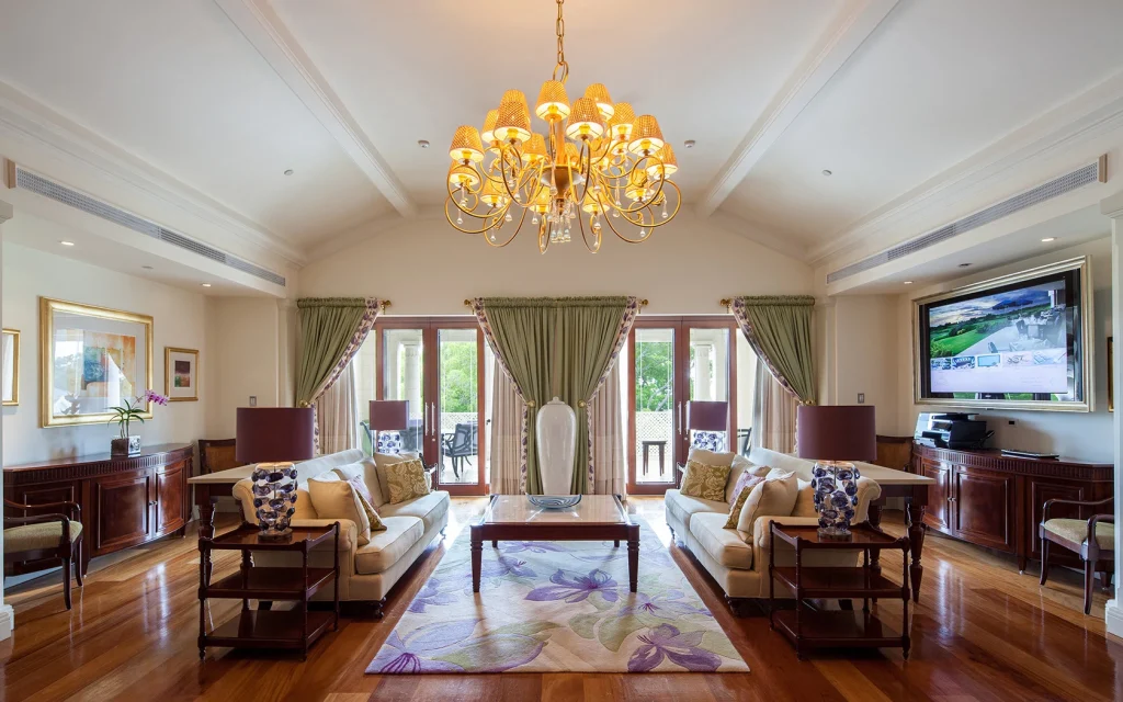 Elegant living room with two beige sofas, floral cushions, a glass coffee table on a floral rug, large chandelier, wooden floors, green curtains on French doors, and a flat-screen TV mounted on the wall.