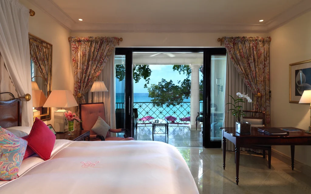 Elegant hotel room with a large bed, floral curtains, a desk, and a balcony overlooking a blue ocean and lush greenery. Soft lighting and tasteful decor create a relaxing, inviting atmosphere.