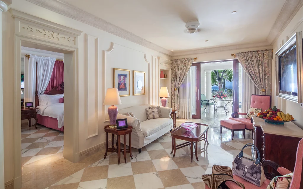 Elegant hotel suite with a bright living area, classic furnishings, and a view of a patio and garden. A bedroom is visible through an open doorway, and sunlight streams in through glass doors. Fresh fruit and handbags are on the tables.