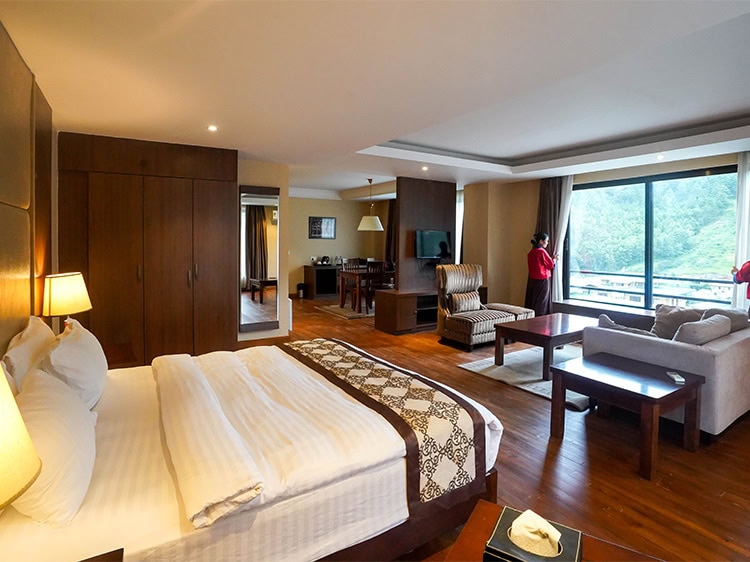 Spacious hotel room with a large bed, couch, armchair, desk, TV, and a big window overlooking greenery. A person stands near the window, and the room has warm lighting and wooden floors.