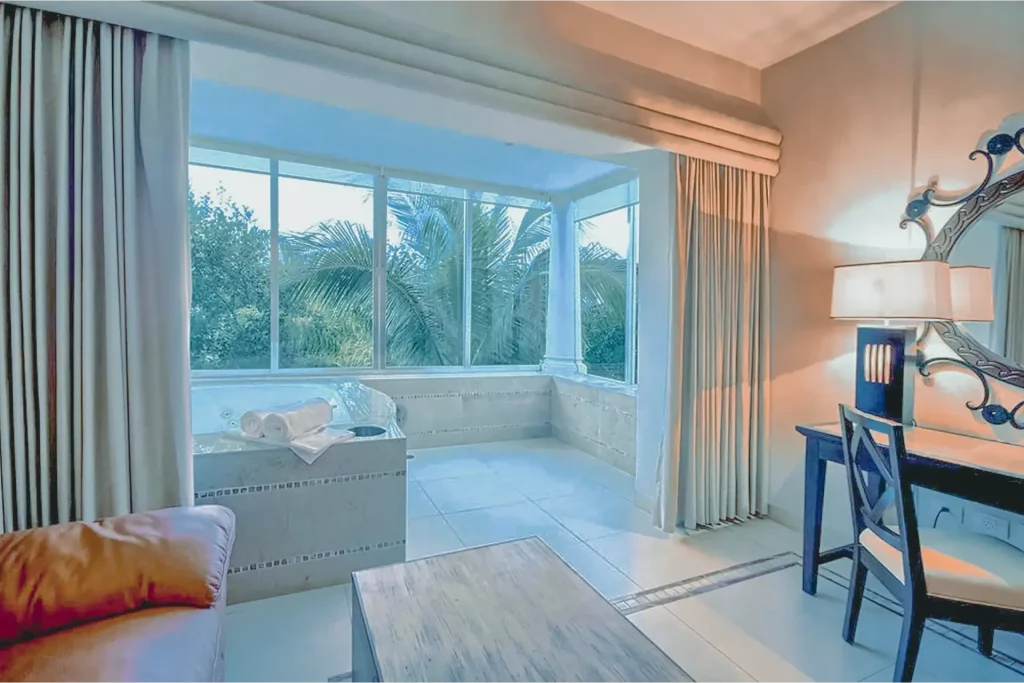 A bright hotel room with a large window, a corner bathtub with towels, beige curtains, a desk with a mirror and a lamp, and a wooden coffee table in front of a sofa. Palm trees are visible outside.