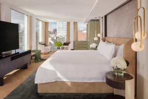 A modern hotel room with a large bed, white linens, bedside tables with white flowers, a flat-screen TV, armchairs by large windows, and city buildings visible outside.