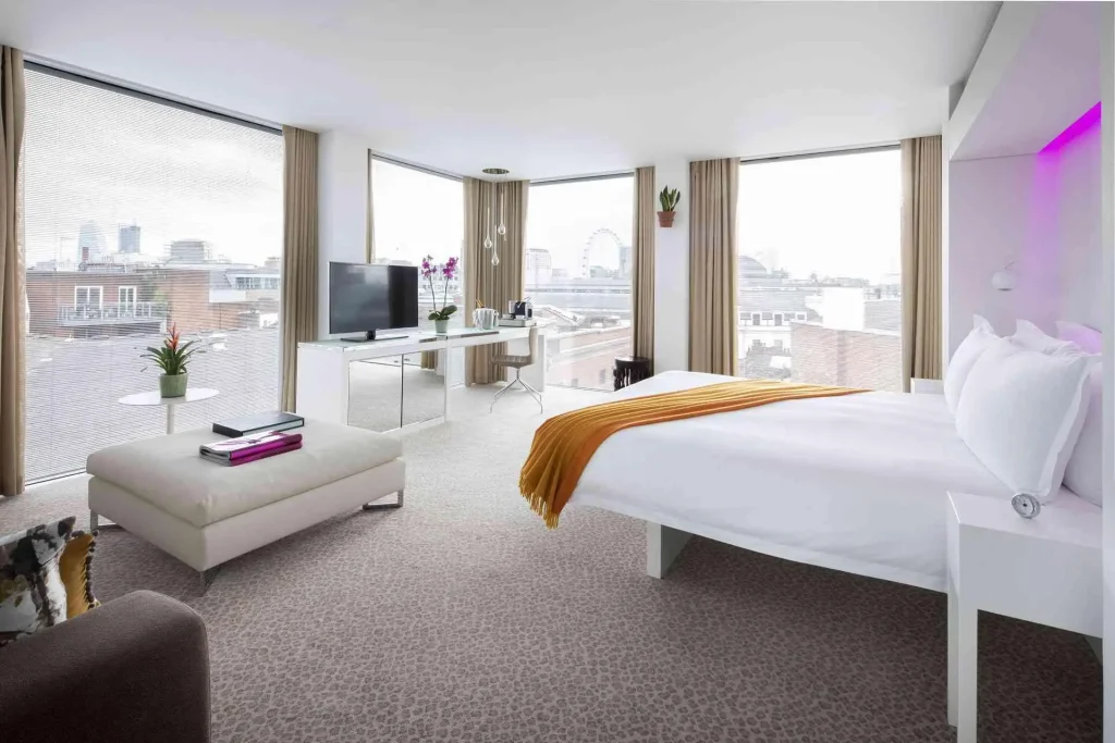 A bright, modern hotel room with large windows, a double bed with a yellow throw, a white ottoman, a desk with a TV, and city views in the background. The room features neutral tones and contemporary decor.