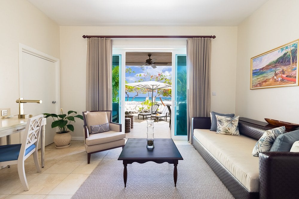 Bright, airy living room with modern furniture, desk, and plant, opening onto a patio with lounge chairs and umbrella overlooking a tropical garden and the ocean in the background.