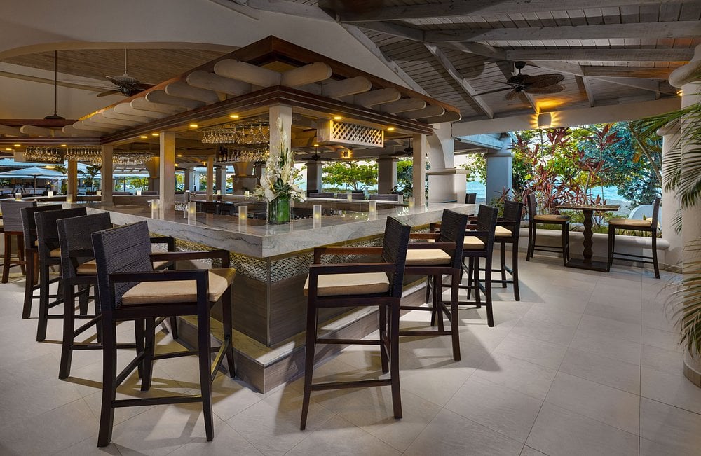 A modern, open-air bar with high chairs around a square marble counter, surrounded by tropical plants and natural light. The space has wooden beams, ceiling fans, and elegant, contemporary decor.