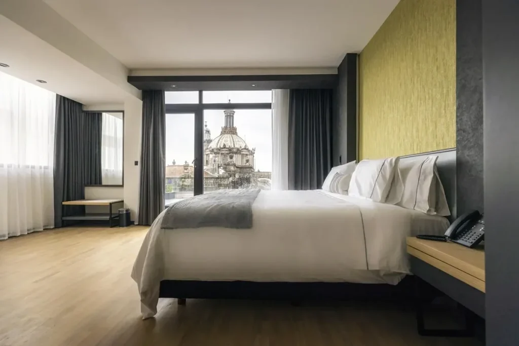 A modern hotel room with a large bed, wooden floor, desk, and floor-to-ceiling windows showing a cityscape with a domed historic building outside.
