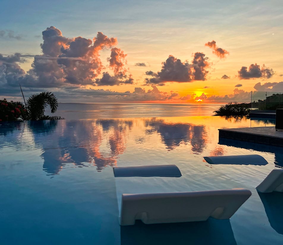 Infinity pool with two white loungers, reflecting a vibrant sunset over a calm ocean. Clouds are illuminated with orange and pink hues, and lush greenery borders the poolside.