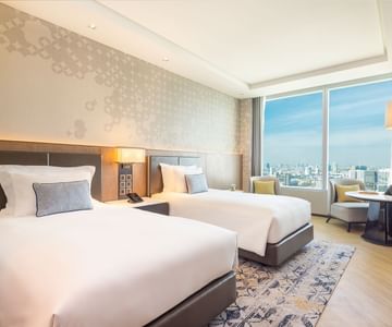 A bright, modern hotel room with two neatly made double beds, a large window offering a city view, a patterned rug, a small table with chairs, and soft, neutral-toned decor.
