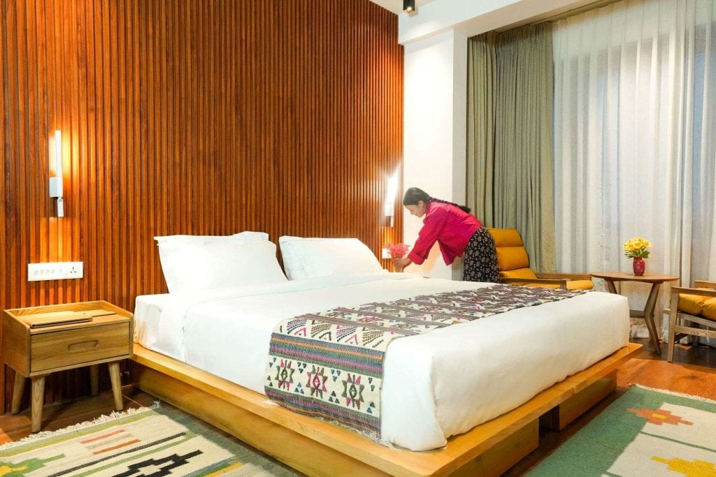 A woman in a pink shirt is making a bed in a modern, well-lit hotel room with wooden panels, white bedding, and patterned decor. There is a nightstand, armchair, small table, and large window with curtains.