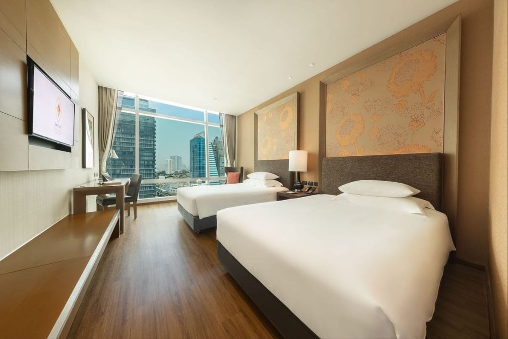 A modern hotel room with two large beds, a wooden floor, a desk and chair by a wide window offering a city view, a TV mounted on the wall, and neutral-colored decor with floral accents.