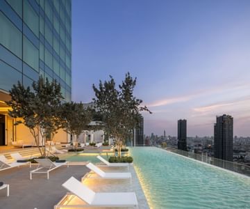 A modern rooftop infinity pool with white lounge chairs and small trees, overlooking a city skyline at sunset. The pool and building are illuminated with soft lights, creating a serene, luxurious atmosphere.