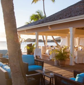 A seaside restaurant patio with wicker chairs, blue cushions, and white tablecloths sits under a shaded roof. Palm trees and ocean views are in the background, creating a relaxing tropical atmosphere.