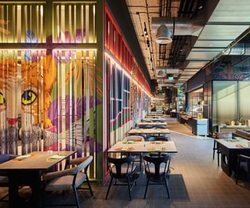 Modern restaurant interior with a vibrant mural of a cat’s face on a partition wall, colorful lighting, neatly arranged tables and chairs, and an open kitchen area visible in the background.