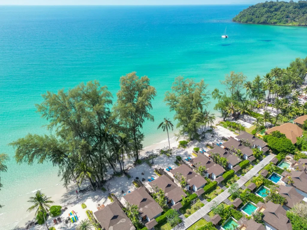 Aerial view of beachfront villas with pools, lush palm trees, and clear turquoise water. A boat floats offshore, and a forested island is visible in the distance under a sunny sky.