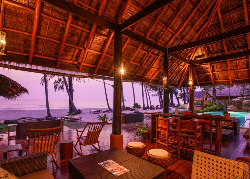 A cozy open-air beachside lounge with wooden furniture and a thatched roof, warmly lit at sunset. The ocean, palm trees, and lounge chairs are visible in the background.
