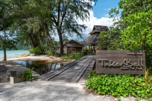 A tropical resort entrance with wooden bridge, lush greenery, palm trees, and a large sign reading Tinkerbell Resort Koh Kood. Huts and a sandy beach with turquoise water are visible in the background.