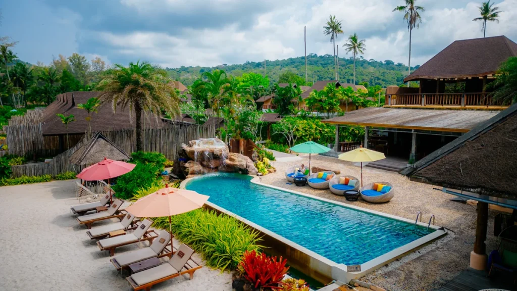 A tropical resort with a rectangular pool, sun loungers, colorful umbrellas, and lush greenery. Thatched-roof buildings and palm trees surround the area, with a sandy space and hills in the background under a cloudy sky.