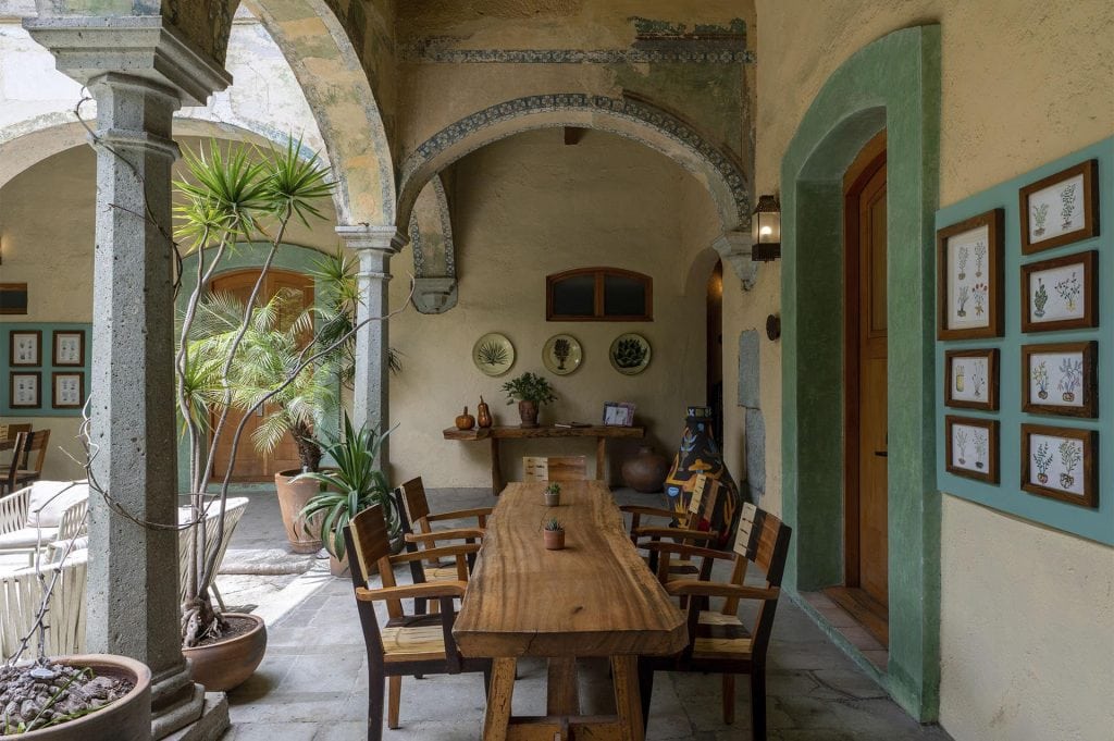 A rustic courtyard with a long wooden table and chairs, surrounded by potted plants and stone arches. Botanical prints hang on the walls, and natural light brightens the cozy, inviting space.