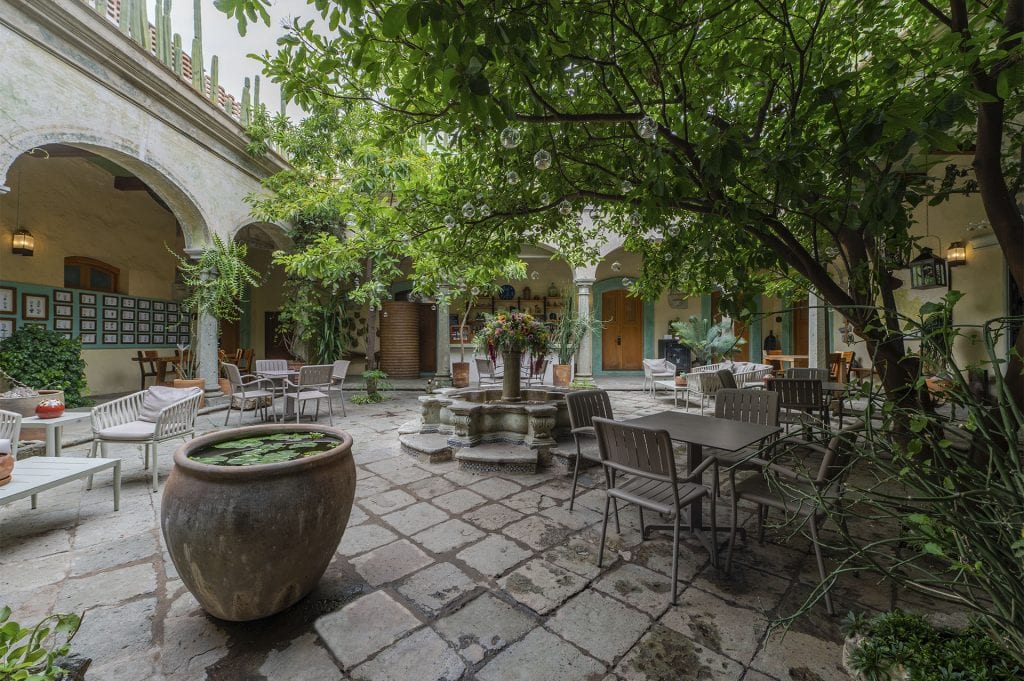 A leafy courtyard with stone flooring, a central fountain, potted plants, and outdoor tables and chairs. The area is surrounded by arched walkways and green doors, creating a tranquil, shaded atmosphere.