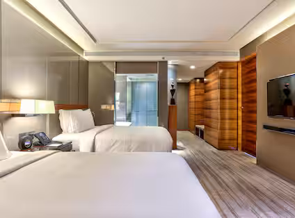 Modern hotel room with two neatly made beds, a wall-mounted TV, wooden accents, bedside lamps, and a glass-walled bathroom in the background. The room features a neutral color palette and contemporary design.