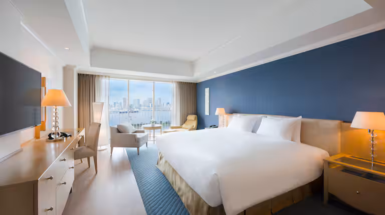 A modern hotel room with a large bed, blue accent wall, two bedside lamps, a desk, armchairs, and floor-to-ceiling windows offering a city skyline and waterfront view.
