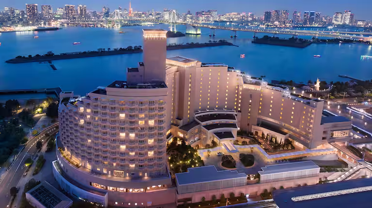 A large, modern hotel with curved architecture is illuminated at night, overlooking a calm bay with a city skyline and a lit bridge in the background. Boats are visible on the water.