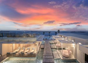 A modern seaside resort at sunset with cabanas, lounge chairs, and pools, overlooking the ocean under a vibrant pink and orange sky.
