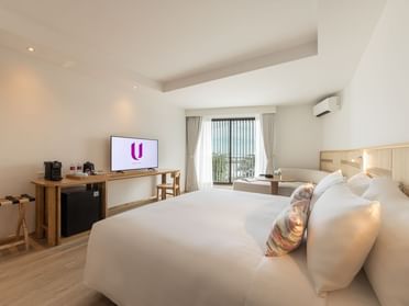 A modern hotel room with a large bed, a TV on a wooden desk, a mini fridge, a coffee station, and a window with curtains letting in natural light, overlooking a balcony with a distant view.