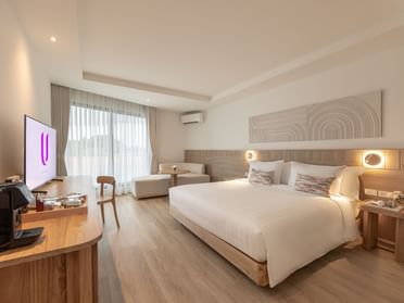 A modern hotel room with a large bed, a wooden desk, a TV, a coffee maker, a small sofa, and floor-to-ceiling windows with sheer curtains letting in natural light. The decor is minimalist with neutral tones.