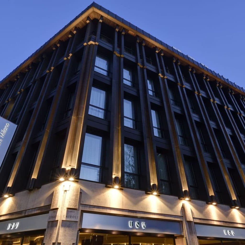 A modern, multi-story building with large windows and illuminated exterior lights at dusk, viewed from a low angle. The signage at the corner reads UNA, indicating its a hotel or commercial property.