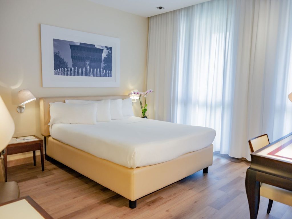 A modern hotel room with a large bed, white linens, light-colored walls, wooden floor, a framed photo above the bed, two wall lamps, a vase with flowers, and sheer curtains over a tall window.