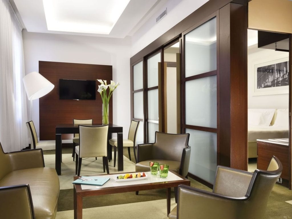 Modern hotel suite with beige and brown furniture, a dining table with flowers, a wall-mounted TV, and a sitting area with a coffee table holding fruit and drinks. Bedroom visible through sliding glass doors.