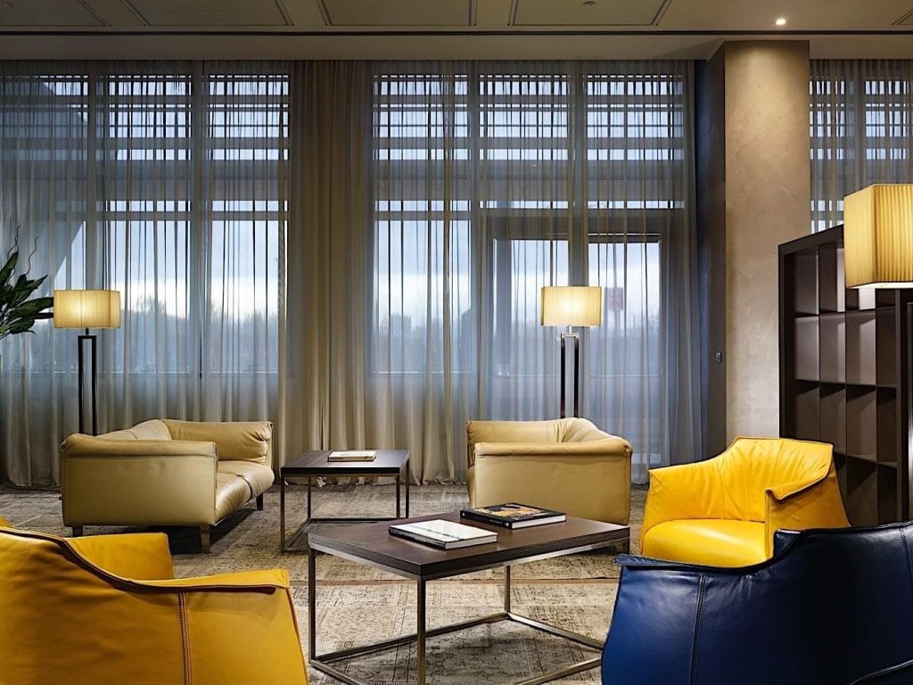 A modern lounge area with large windows, sheer curtains, cozy beige and yellow armchairs, dark coffee tables with books, soft lighting from floor lamps, and a contemporary, relaxed atmosphere.