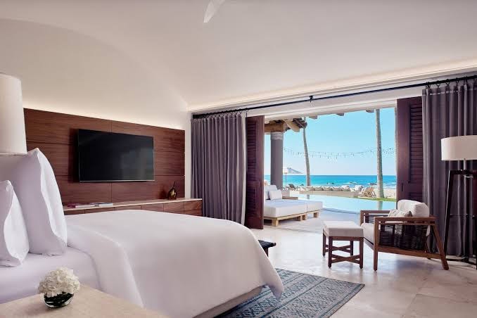 A modern hotel room with a large bed, a flat-screen TV, and seating area. Sliding doors open to a terrace with lounge chairs and a pool, overlooking the ocean under a sunny sky.