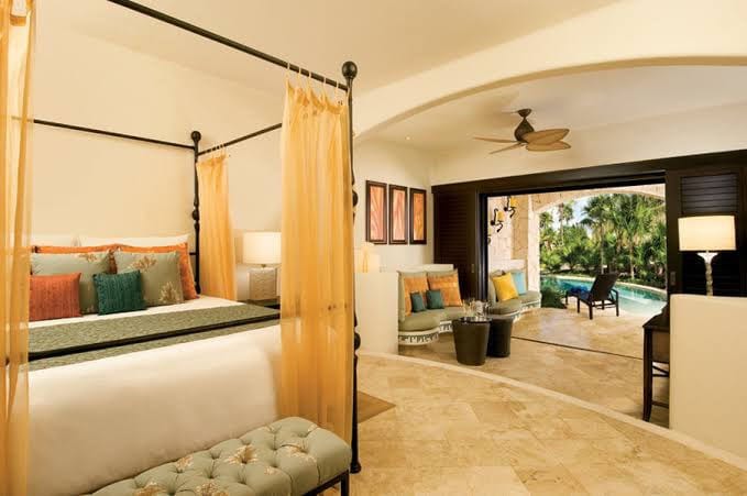 Luxurious hotel room with a four-poster bed, plush seating area, tropical decor, and open doors leading to a patio with a pool and lush garden view. Warm lighting creates an inviting atmosphere.
