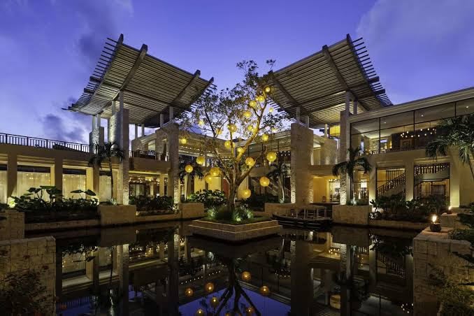 A luxurious resort building with open, modern architecture, lit warmly at dusk. A central tree decorated with glowing lights stands above a reflective pool surrounded by stone columns and lush greenery.