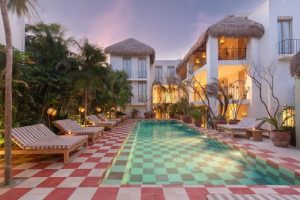 A rectangular pool with a checkered tile deck is surrounded by lounge chairs, tropical plants, and a two-story building with thatched roofs at sunset. Warm lights create a cozy ambiance.