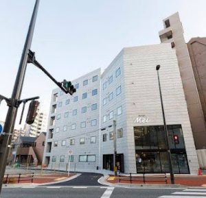 A modern, multi-story white building labeled Hotel Mei stands at a street corner with traffic lights and crosswalks on a clear day.