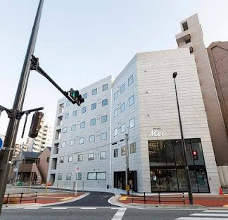 A modern, multi-story white building labeled Hotel Mei stands at a street corner with traffic lights and crosswalks on a clear day.