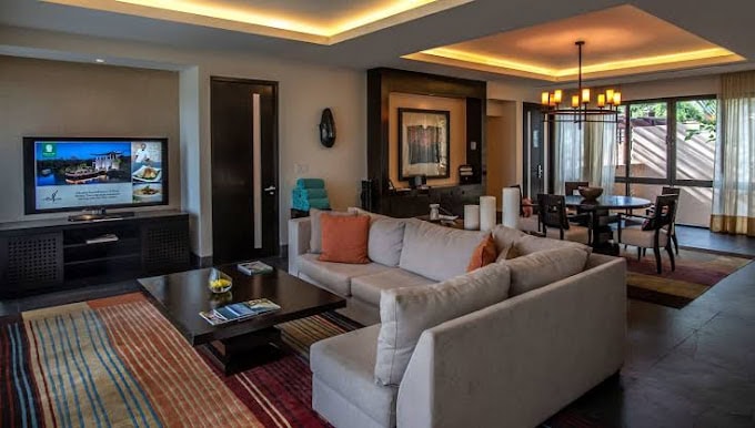 Modern living room with a beige sectional sofa, colorful pillows, and striped rug, facing a TV. Dining area with a chandelier and large window is visible in the background. Warm lighting creates a cozy atmosphere.