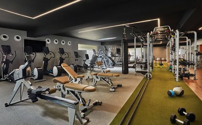 Modern gym with various exercise machines, free weights, benches, rowing machines, and cross trainers. There is a turf strip with medicine balls and battle ropes, under bright lighting and dark ceiling.