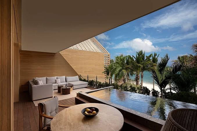 A luxurious terrace features a private plunge pool, a round table with a fruit bowl, and a cozy outdoor sofa, overlooking palm trees and a scenic beach under a partly cloudy sky.