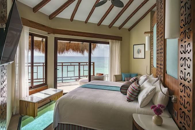 A cozy bedroom with a king-size bed, modern decor, wooden beams, and large windows overlooking a balcony with a stunning ocean view. Light floods the room, creating a relaxing, tropical atmosphere.