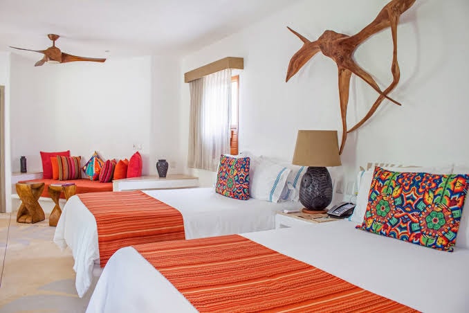 A brightly lit hotel room with two beds, colorful patterned pillows, orange throws, a wooden sculpture on the wall, a lamp, and a small seating area with red and multicolored cushions.