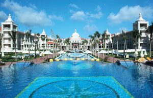 A large, luxurious resort features a wide swimming pool with geometric patterns, surrounded by palm trees, lounge chairs, and grand white buildings under a bright blue sky.