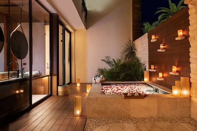 A luxurious outdoor spa area at night, featuring a square hot tub filled with rose petals, surrounded by candles on a wooden wall and floor, adjacent to a modern glass-walled bathroom with lush plants nearby.