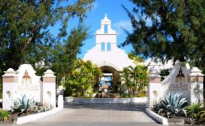 Entrance to Spice Island Beach Resort with white archways, tropical plants, and clear blue sky, flanked by two white signs and lush greenery.